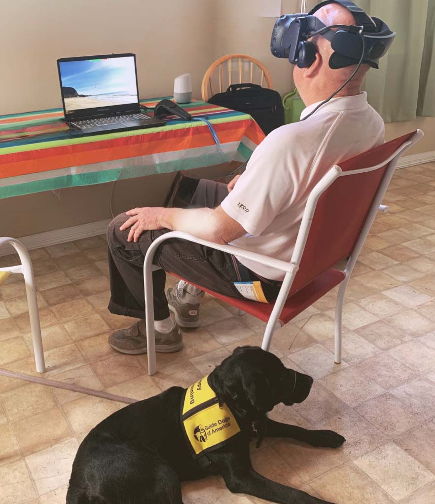 Photo of a man engaging with virtual reality, with his service dog at his side.